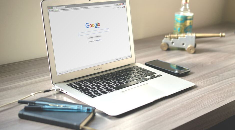 A neat workspace featuring a laptop displaying Google search, a smartphone, and a notebook on a wooden desk.