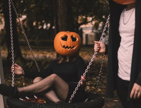 Creative Halloween photo with two people wearing pumpkin masks, one sitting on a swing outdoors.