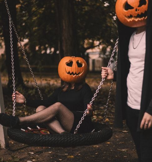 Creative Halloween photo with two people wearing pumpkin masks, one sitting on a swing outdoors.